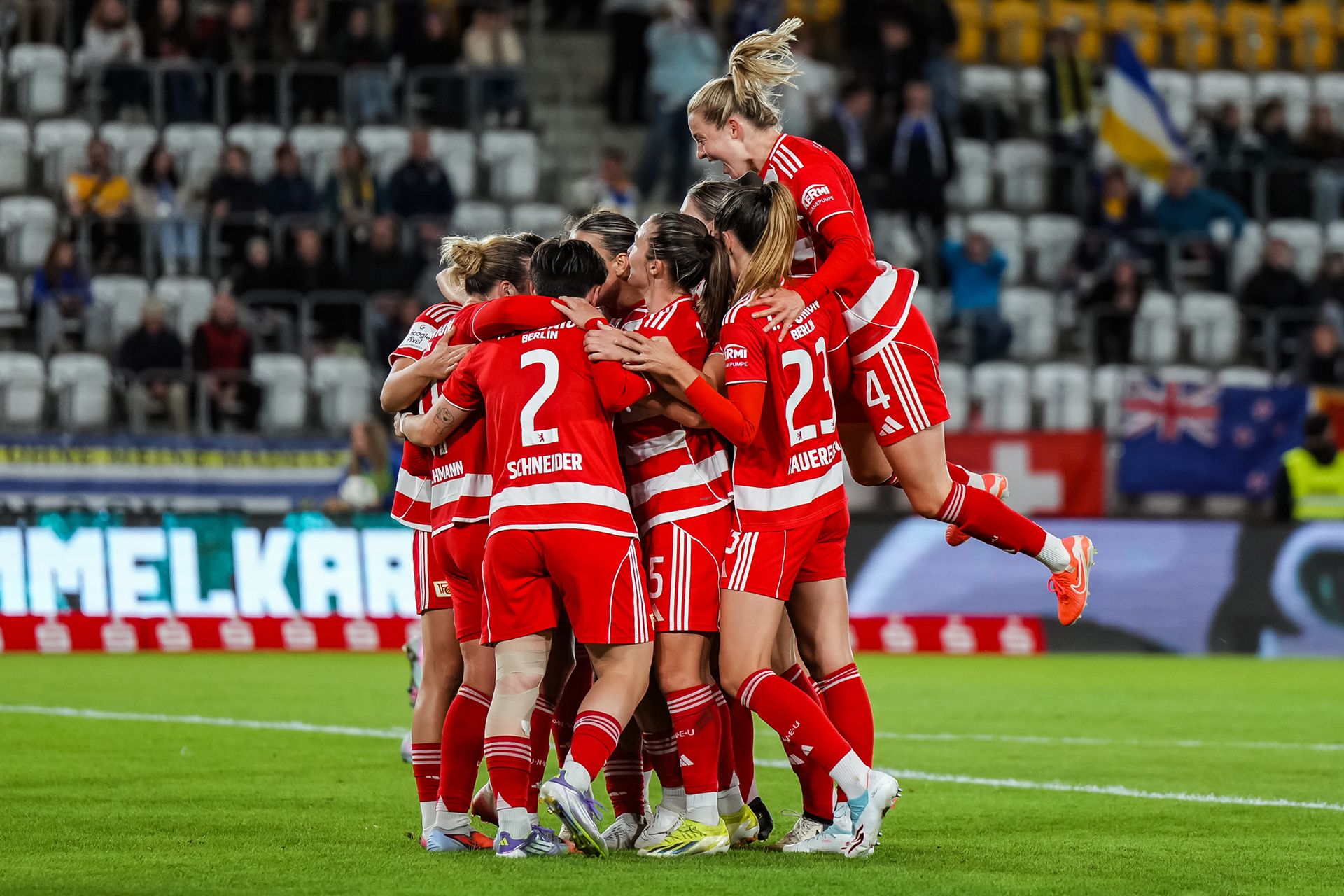 Eine Gruppe von Frauenfußballspielerinnen in roten Trikots feiert einen Torerfolg mit Jubel und Umarmungen auf dem Spielfeld.