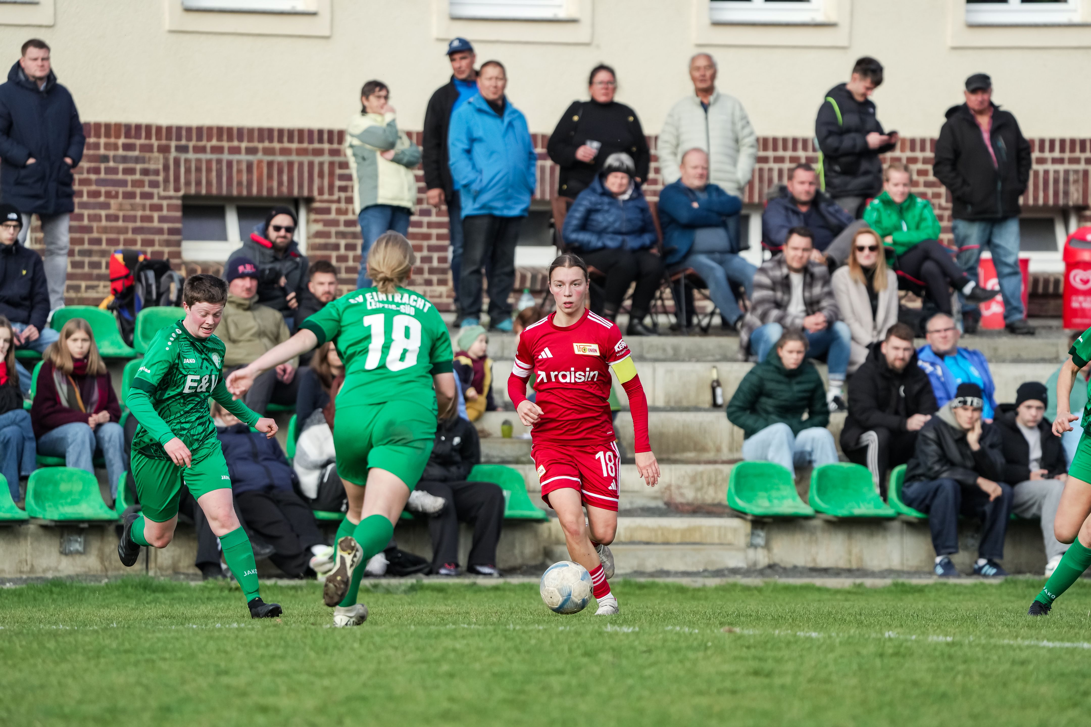 Eine Fußballspielerin in roter Ausrüstung dribbelt den Ball, während eine gegnerische Spielerin in grüner Ausrüstung näherkommt. Zuschauer im Hintergrund.