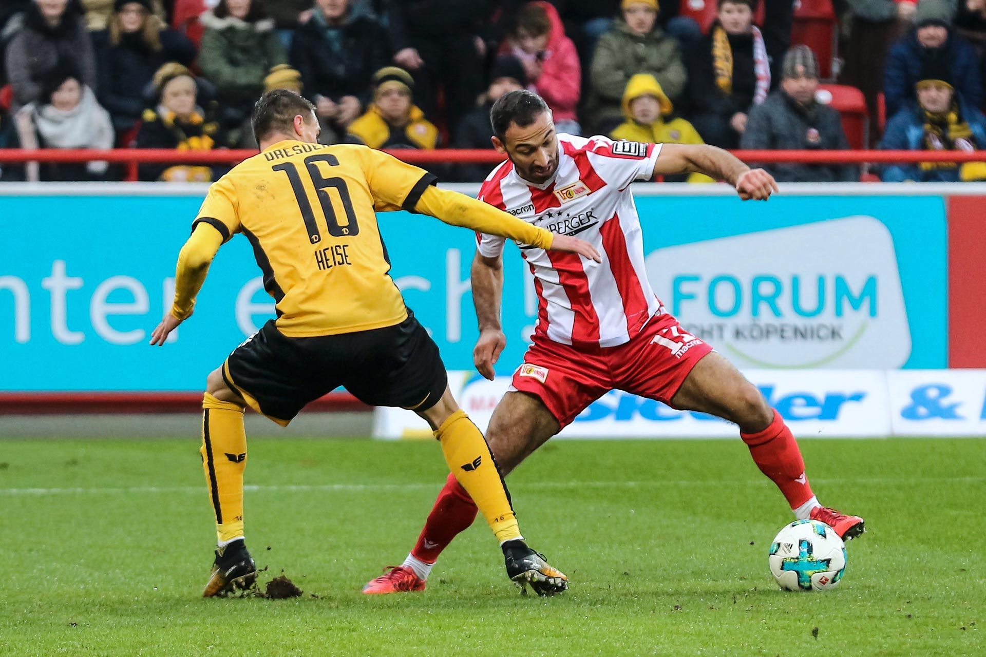 Zwei Spieler kämpfen um den Ball. Viele Zuschauer schauen zu. Es ist eine spannende Stimmung im Stadion.