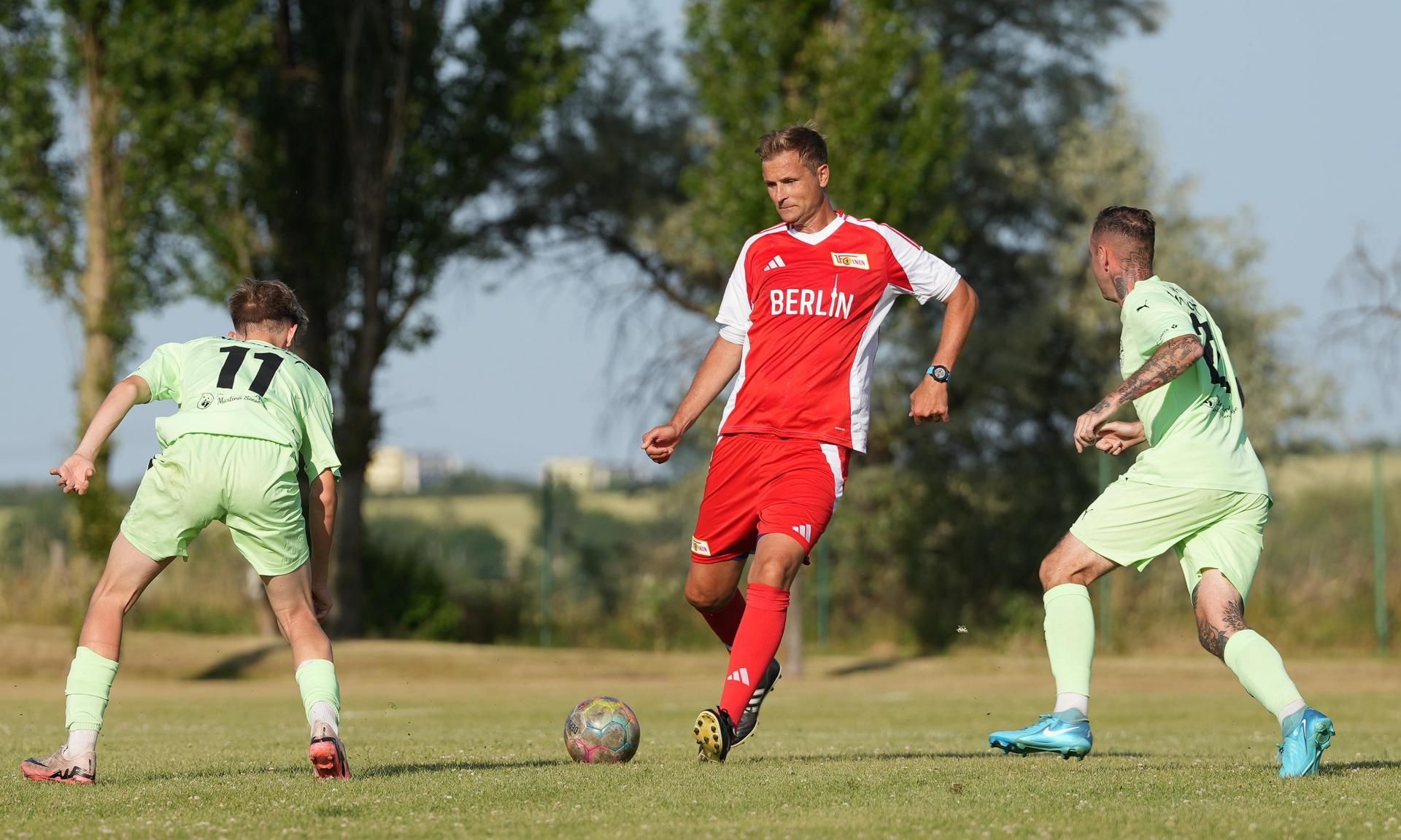 Ein Fußballspieler trägt ein rotes Trikot. Er dribbelt den Ball. Zwei Gegner in grünen Trikots sind in der Nähe. Sie wollen den Ball holen.