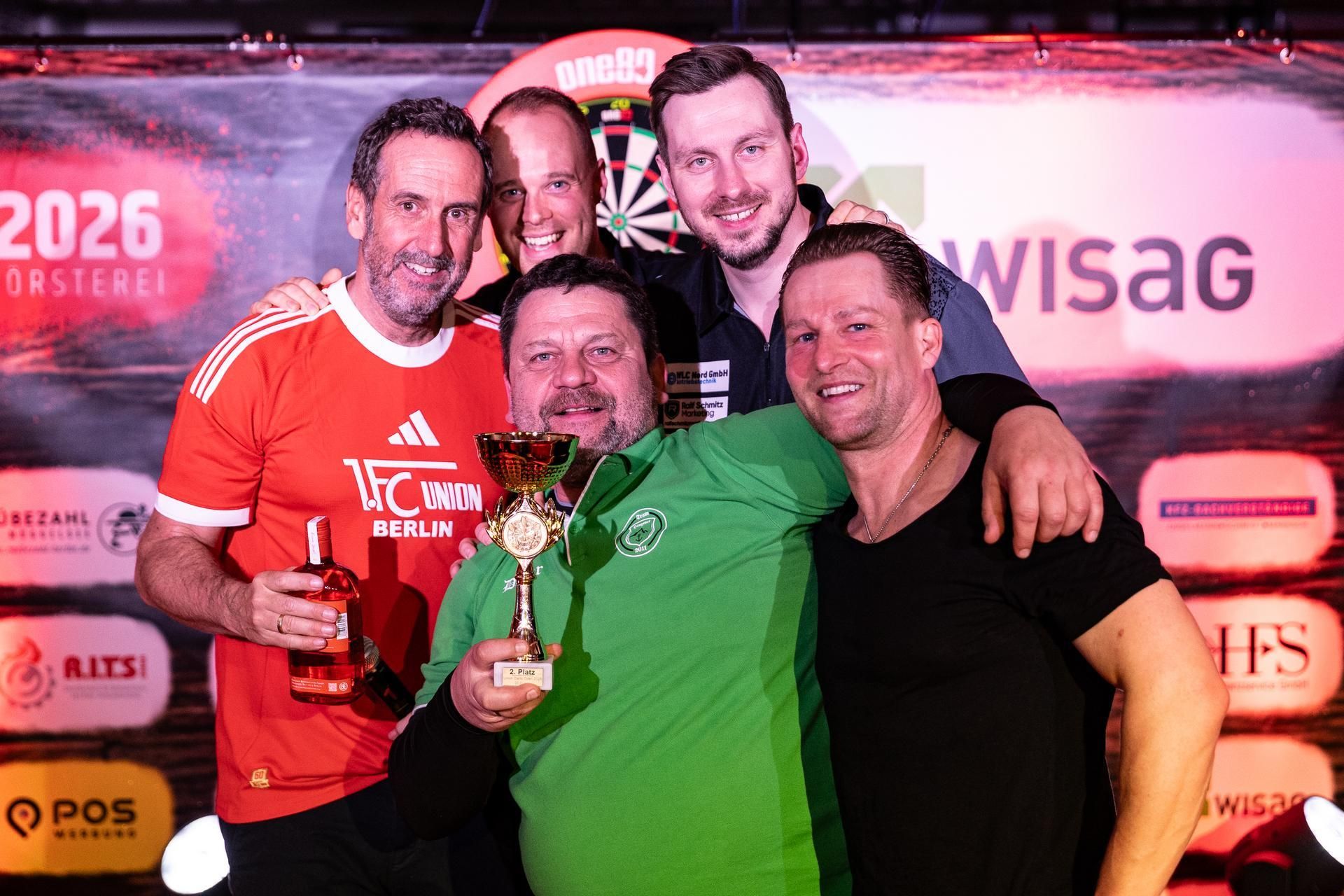 Fünf Männer posieren lächelnd zusammen, einer hält einen Pokal in der Hand, im Hintergrund ein buntes Event-Backdrop.