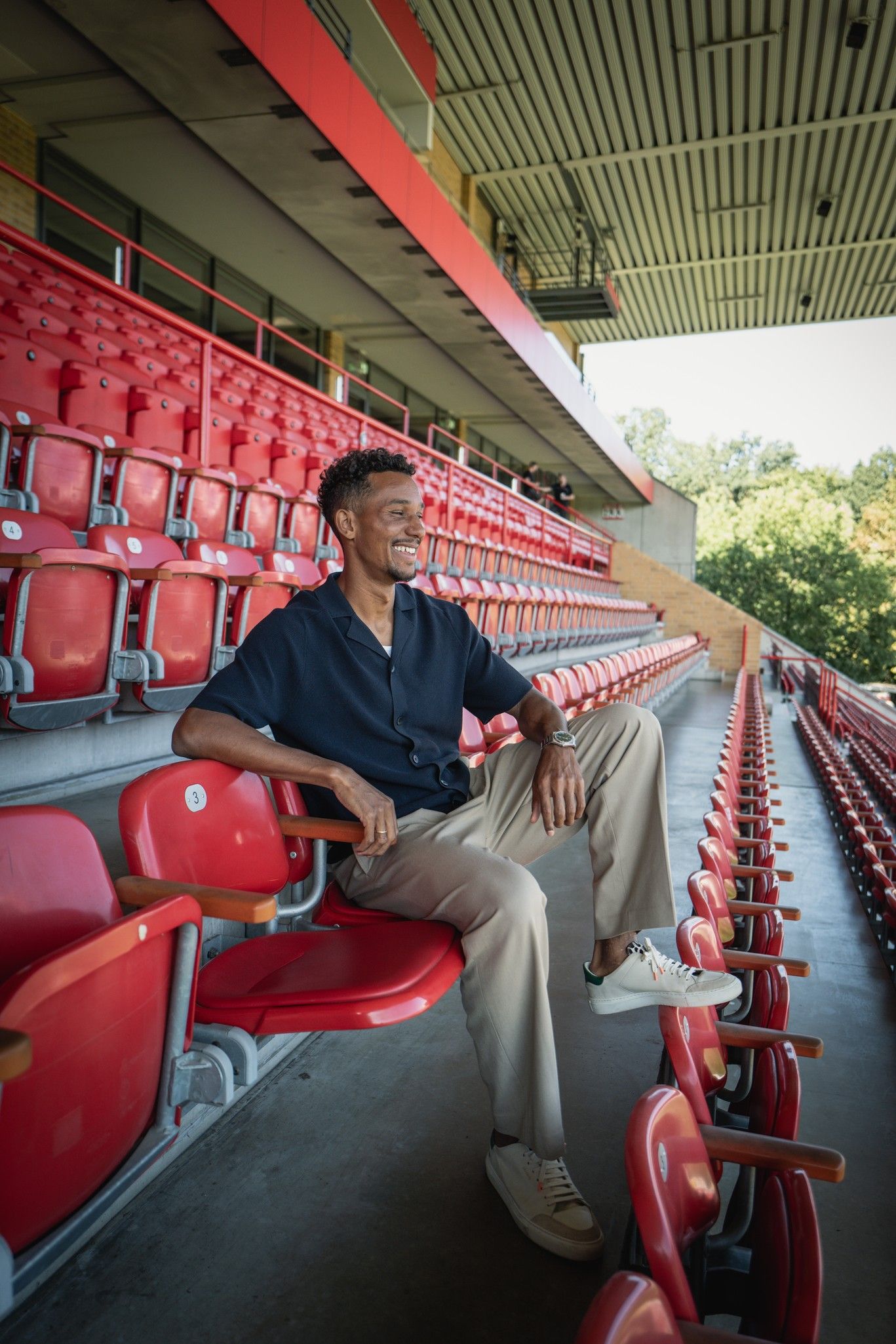 Ein Mann sitzt entspannt auf einem roten Stadionplatz zwischen leeren Sitzen und lächelt.