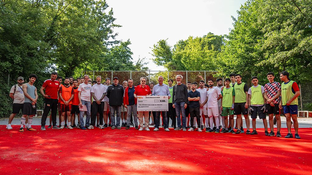 Gruppenszene auf einem roten Sportplatz mit Jugendlichen und Erwachsenen, die gemeinsam einen großen Scheck präsentieren.