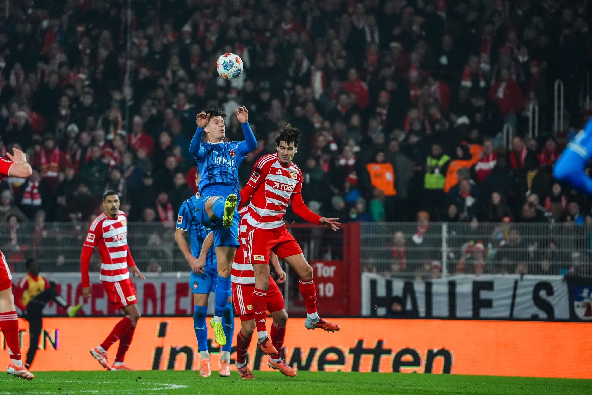 Fußballspiel mit Spielern in roten und blauen Trikots, ein Spieler springt zum Kopfball über einen anderen. Stadionatmosphäre im Hintergrund.