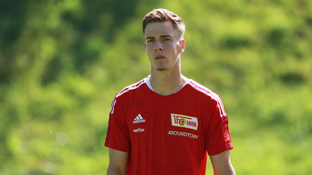 Ein junger Fußballspieler in rotem Trikot steht im Freien, mit unscharfem, grünem Hintergrund.