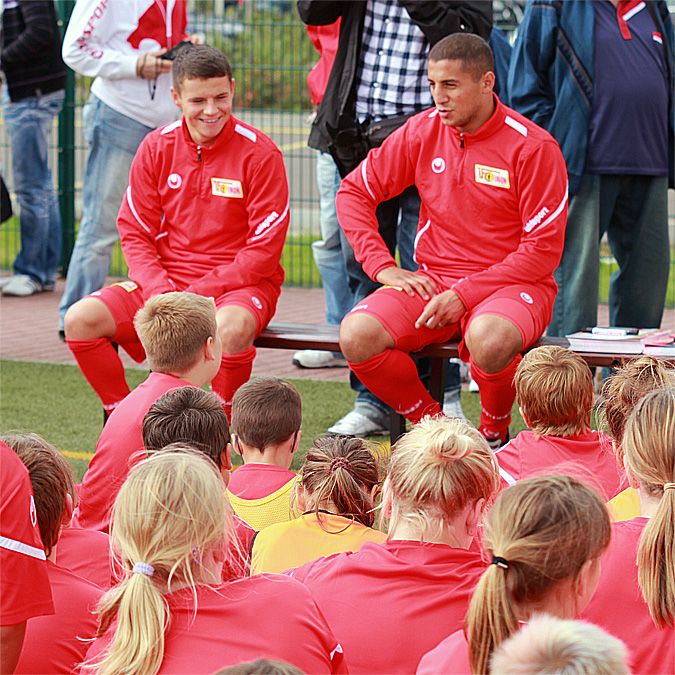 Zwei fußballspielende Trainer in roter Sportkleidung sitzen vor einer Gruppe junger Spieler auf einem Trainingsfeld.
