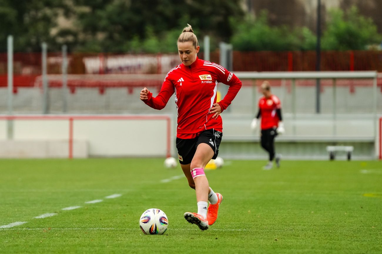 Frau im roten Trainingsshirt tritt einen Fußball auf einem Sportplatz. Im Hintergrund sind weitere Spielerinnen zu sehen.