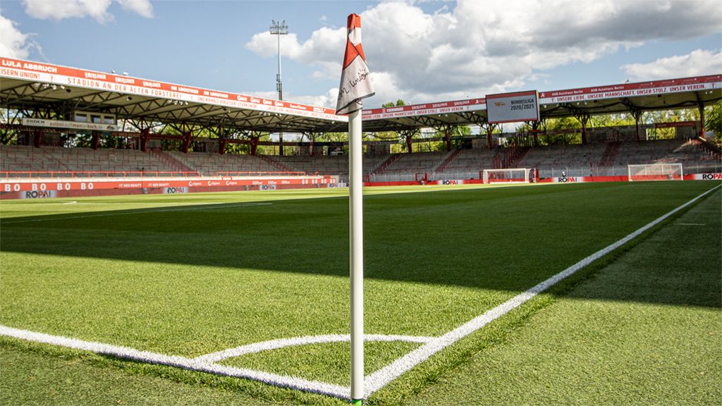 Eckfahne in einem Fußballstadion mit Rasenplatz und Tribünen im Hintergrund, blauem Himmel und Wolken.