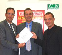 Drei Männer in Anzügen stehen zusammen und halten ein Dokument in der Hand, im Hintergrund ein Werbeplakat.