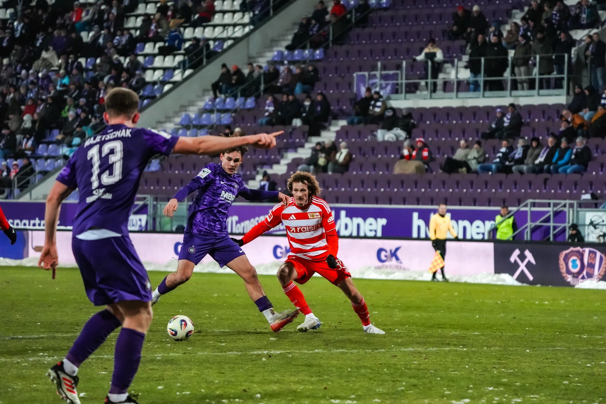 Zwei Fußballspieler stehen sich auf dem Rasen gegenüber, während ein dritter Spieler im Hintergrund das Spiel beobachtet. Stadionatmosphäre.