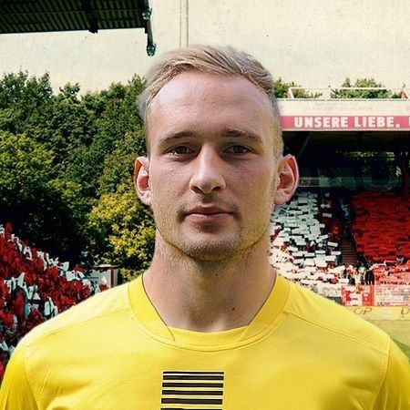 Mann mit kurzem Haar in gelbem Fußballtrikot steht vor einem Stadion mit Zuschauertribünen.