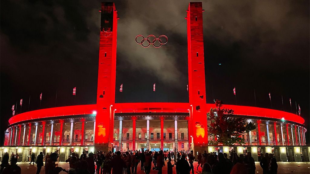 Olympic Stadium at night, illuminated in red, with the five Olympic rings and crowds in the foreground.