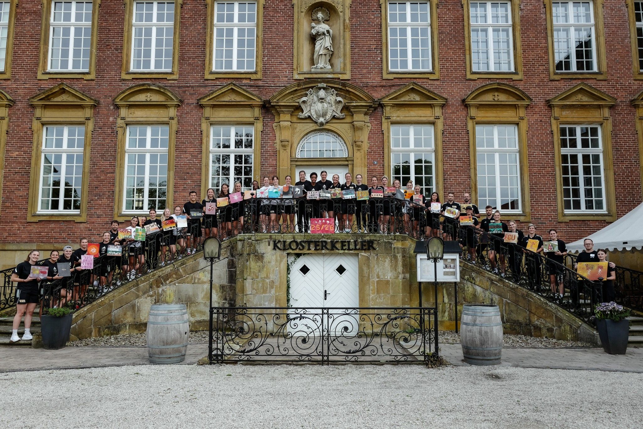 Gruppenfoto von Personen auf einer Treppe vor einem historischen Gebäude, alle halten bunte Plakate in den Händen.