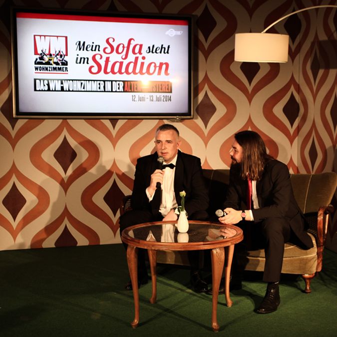 Zwei Männer in Anzügen sitzen auf einem Sofa vor einer großen Wandgrafik mit Veranstaltungshinweisen.