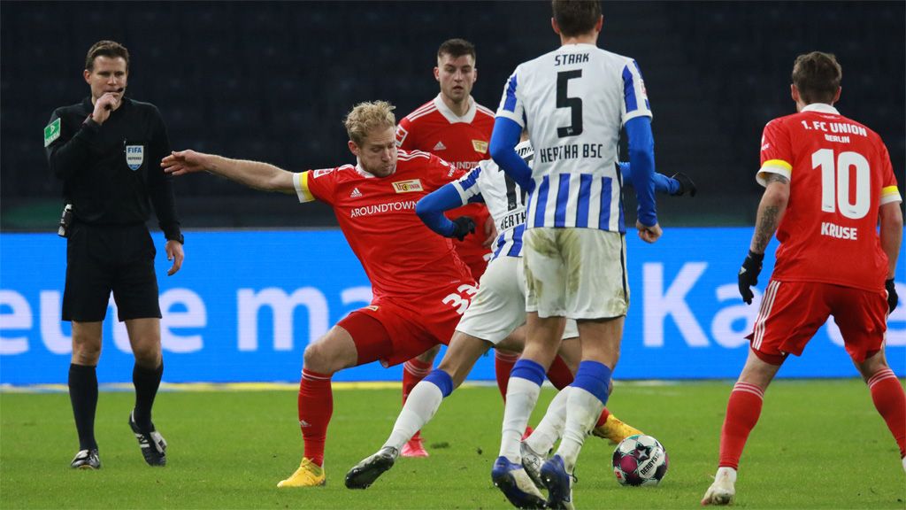 Ein Fußballspiel mit Spielern der Teams in roten und blauen Trikots, ein Spieler fällt zu Boden, Schiedsrichter steht daneben.