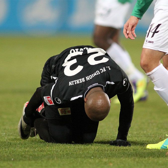 Ein Fußballspieler in schwarzem Trikot kniet am Boden, während ein anderer Spieler in grünem Trikot daneben steht.