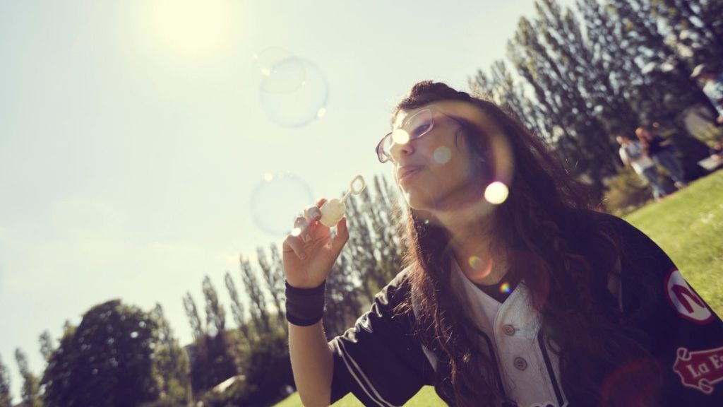 Eine Person bläst Seifenblasen auf einer Wiese, umgeben von Bäume und Sonne, die im Hintergrund scheint.