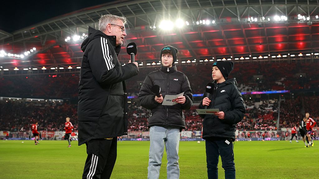 Ein Mann steht mit Mikrofon auf dem Spielfeld und spricht mit zwei Jungen, während im Hintergrund das Stadion beleuchtet ist.
