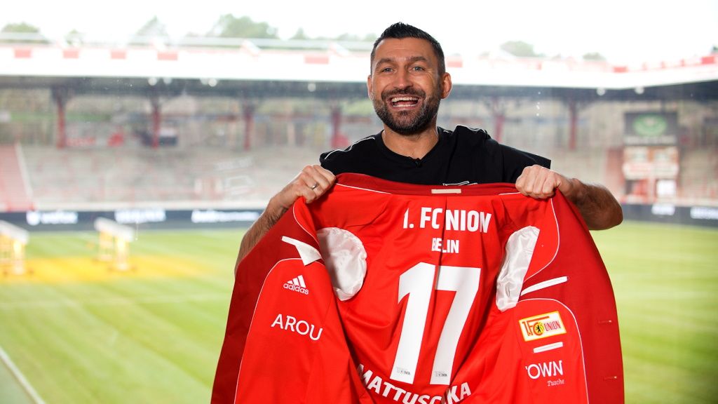A smiling soccer player holds a red jersey with the number 17 and the name "Nattuska" in front of a stadium backdrop.