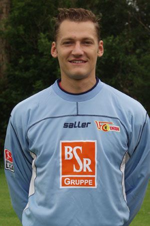Ein Fußballspieler in einem blauen Trainingsanzug steht lächelnd vor einer grünen Hintergrundlandschaft.