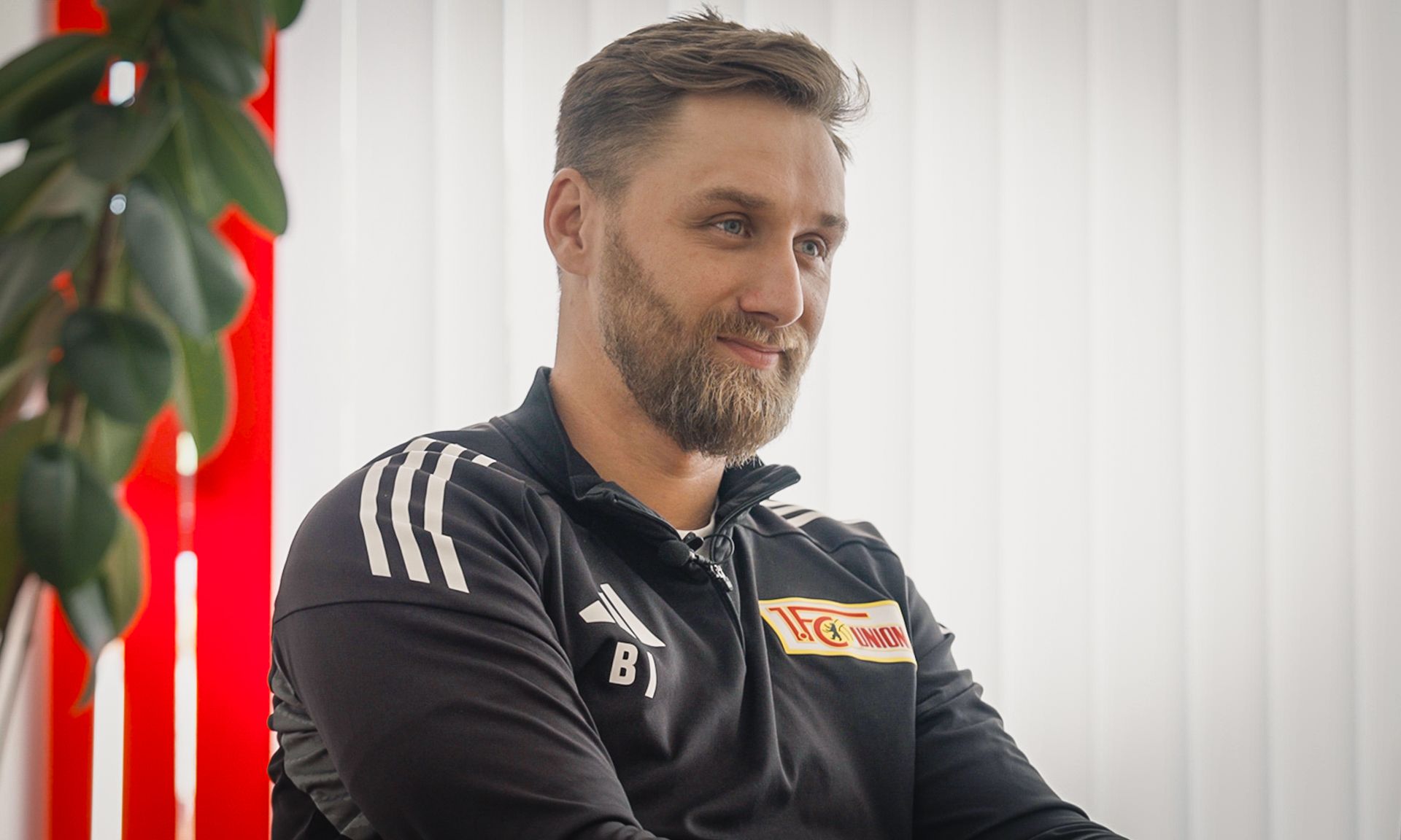 Ein lächelnder Mann mit Bart trägt ein schwarzes Sportoberteil mit einem Vereinslogo. Im Hintergrund sind helle vertikale Jalousien.