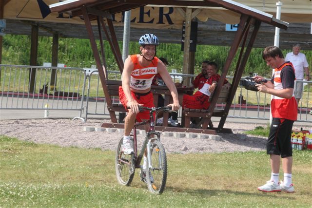 Ein Mann in Sportkleidung auf einem Fahrrad, während ein anderer im Hintergrund mit einer Kamera filmt. Sonnige, grüne Umgebung.