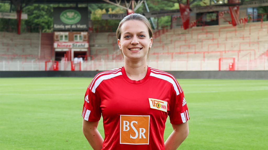 Eine lächelnde Frau in einem roten Fußballtrikot mit dem Logo eines Vereins steht auf einem Stadionrasen.