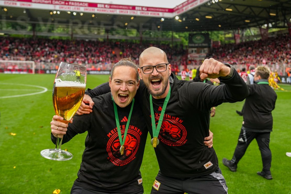 Zwei jubelnde Personen feiern auf einem Fußballplatz, halten ein großes Bierglas und tragen Medaillen. Hintergrund: jubelnde Menge.