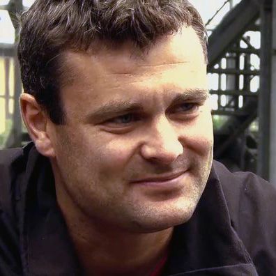 Ein Mann mit dunklen Haaren und einem freundlichen Lächeln sitzt in einer urbanen Umgebung.