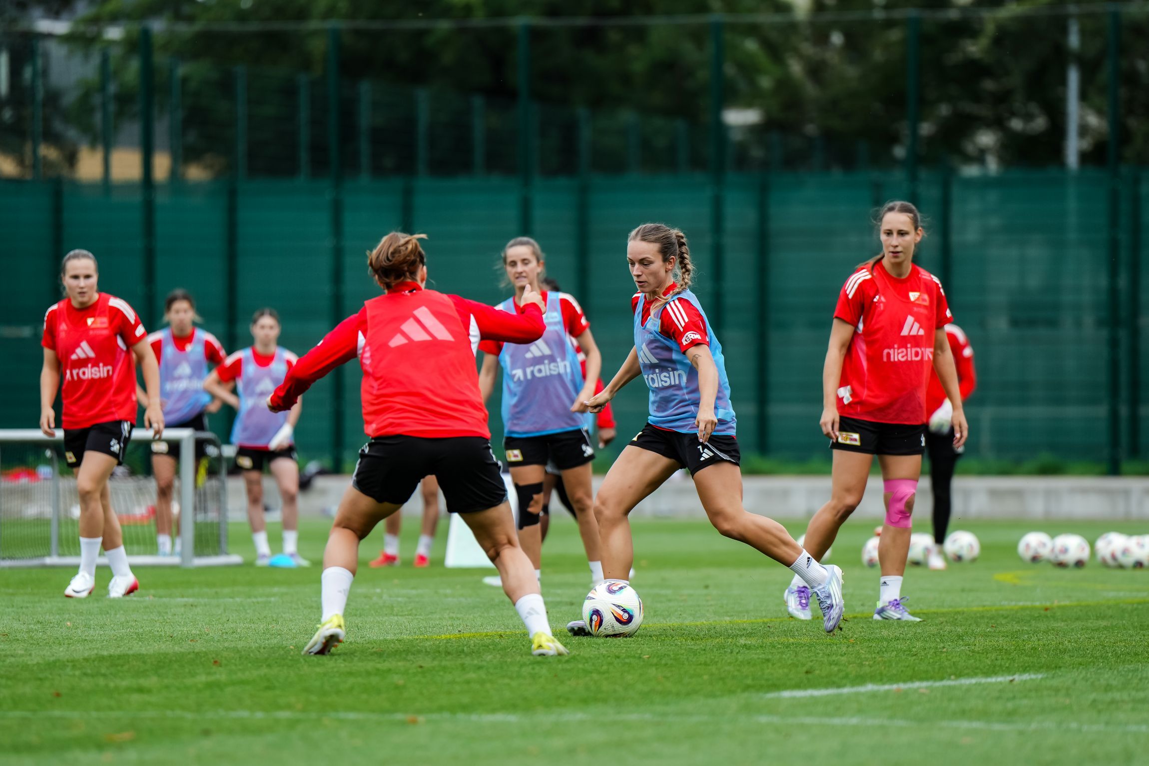 Eine Gruppe von Spielerinnen trainiert auf einem Fußballfeld, während eine Spielerin den Ball dribbelt und eine andere sie im Spiel verfolgt.