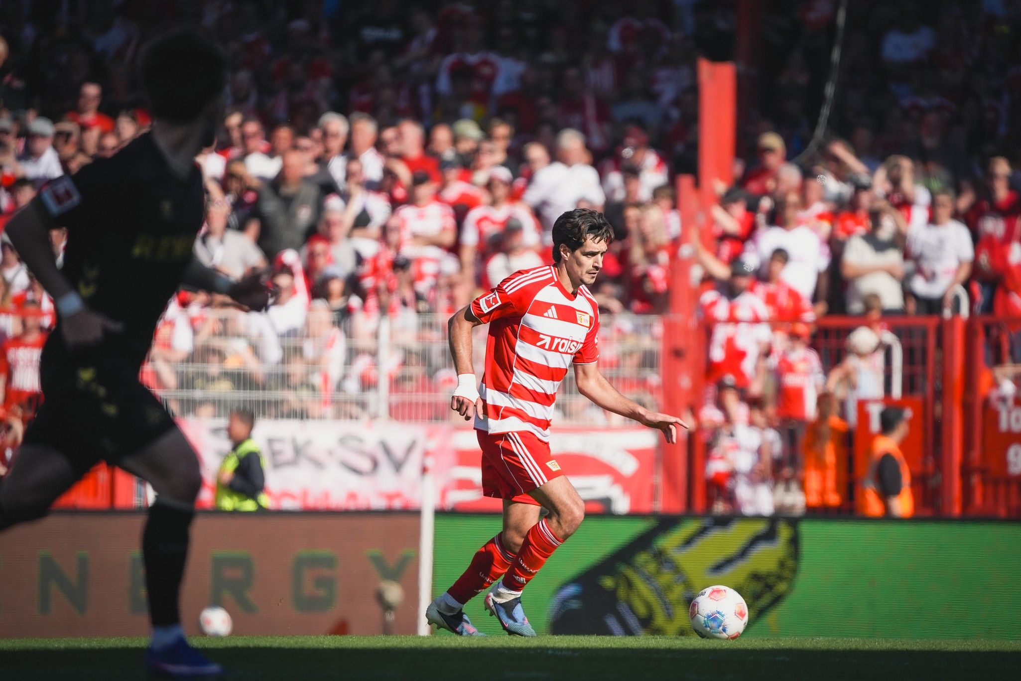 Spielszene eines Fußballmatches mit einem Spieler in rotem Trikot, umgeben von jubelnden Fans im Stadion.