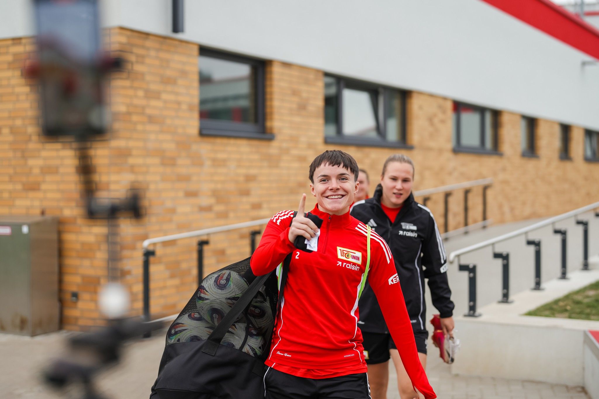 Junge in rotem Sportoutfit mit Tasche, lächelt zur Kamera, während eine weitere Person in sportlicher Kleidung neben ihm geht.