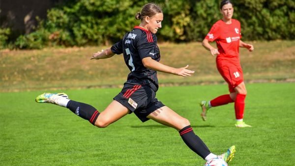 A female soccer player in a black jersey takes a shot while an opponent in a red jersey watches.