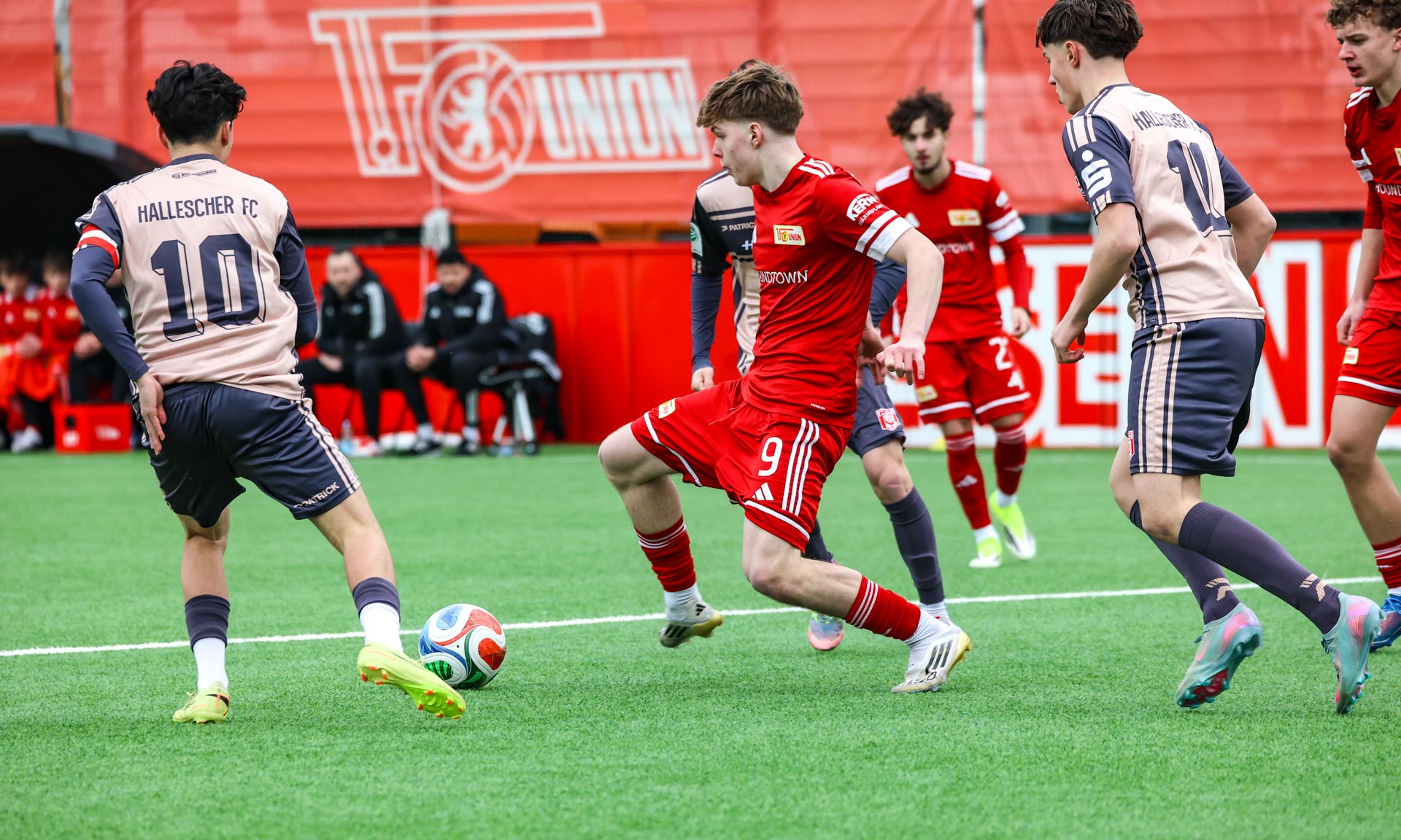 Ein Fußballspiel auf einem Kunstrasenplatz, Spieler in roten und grauen Trikots kämpfen um den Ball.
