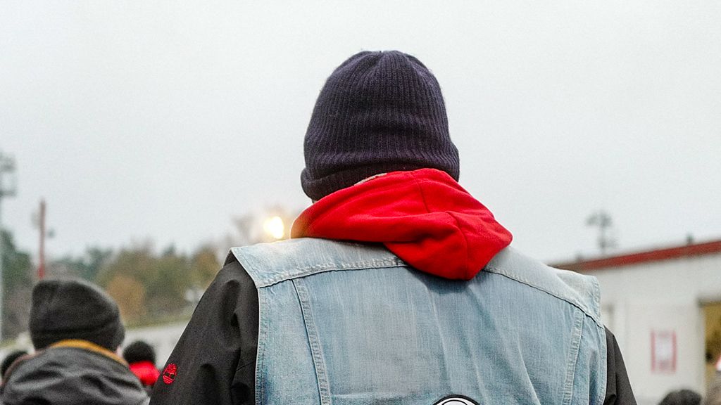 Person from behind, wearing a gray denim jacket and a red hoodie, with a dark beanie on their head. Cloudy sky.
