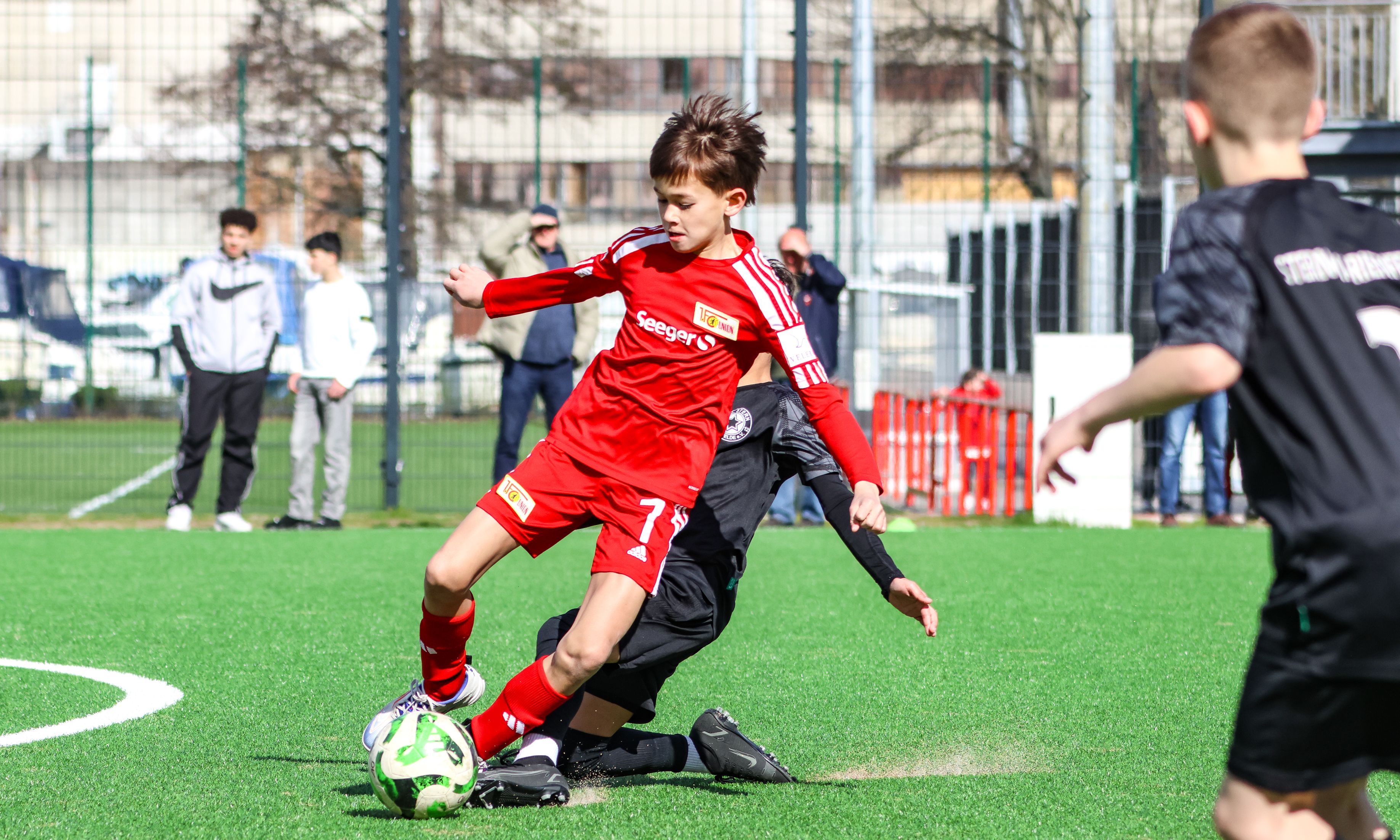 Ein junger Fußballspieler trägt ein rotes Trikot. Er dribbelt den Ball. Ein anderer Spieler ist nah bei ihm.