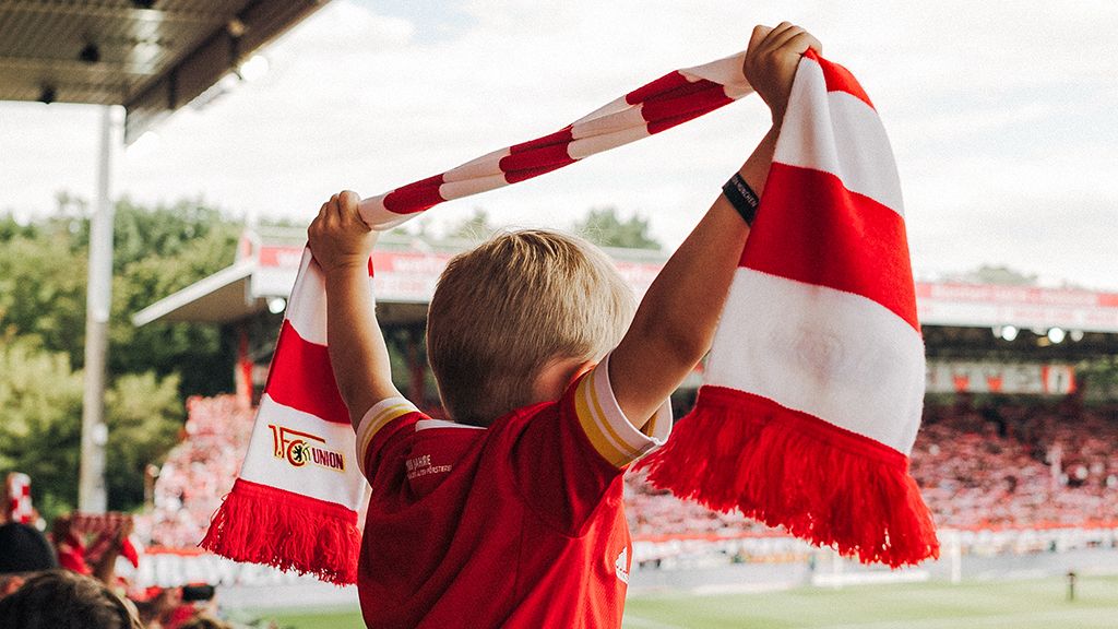 Ein Junge in einem roten Trikot hält einen rot-weißen Schal hoch und jubelt in einem Stadion voller Fans.