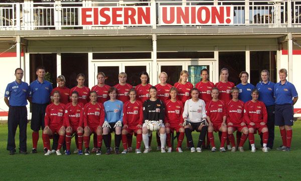 Gruppenfoto der Frauenmannschaft von Eisern Union in roten Trikots auf einem Sportplatz, mit Trainern und Spielerinnen.