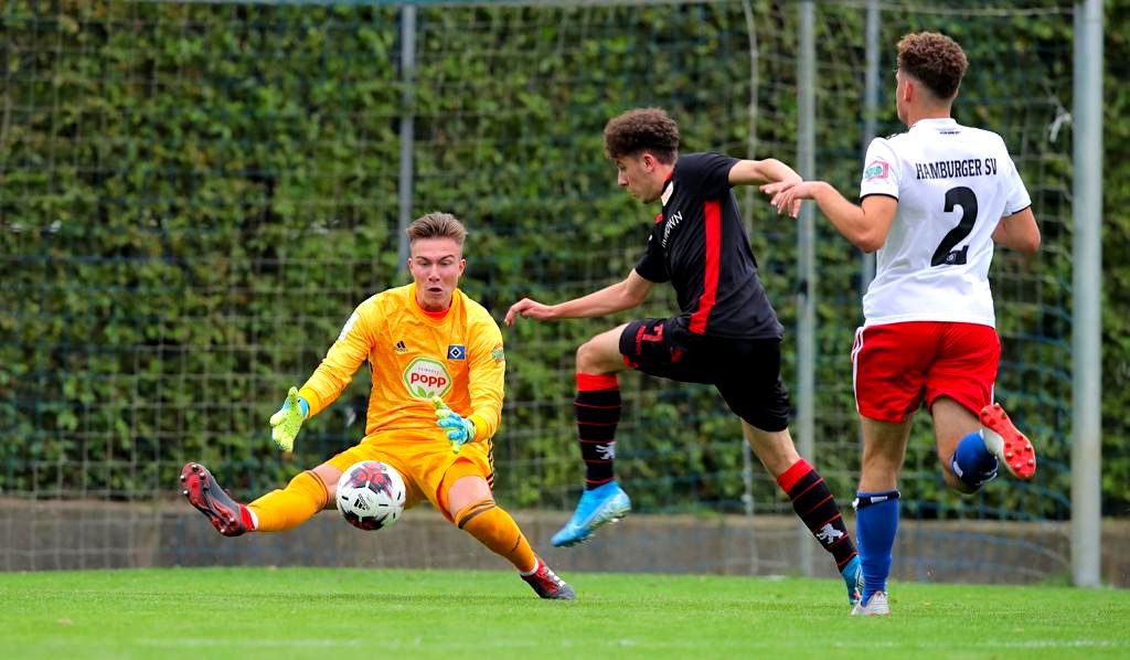 Ein Torwart in gelb wehrt einen Schuss ab, während ein angreifender Spieler in schwarz und ein verteidigender Spieler in weiß agieren.