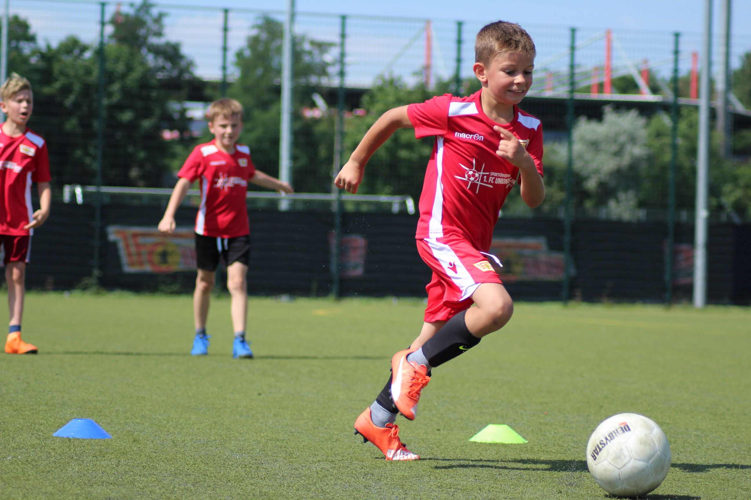 Ein junger Junge in rotem Trikot läuft mit einem Fußball über ein Trainingsfeld, während zwei weitere Kinder im Hintergrund üben.