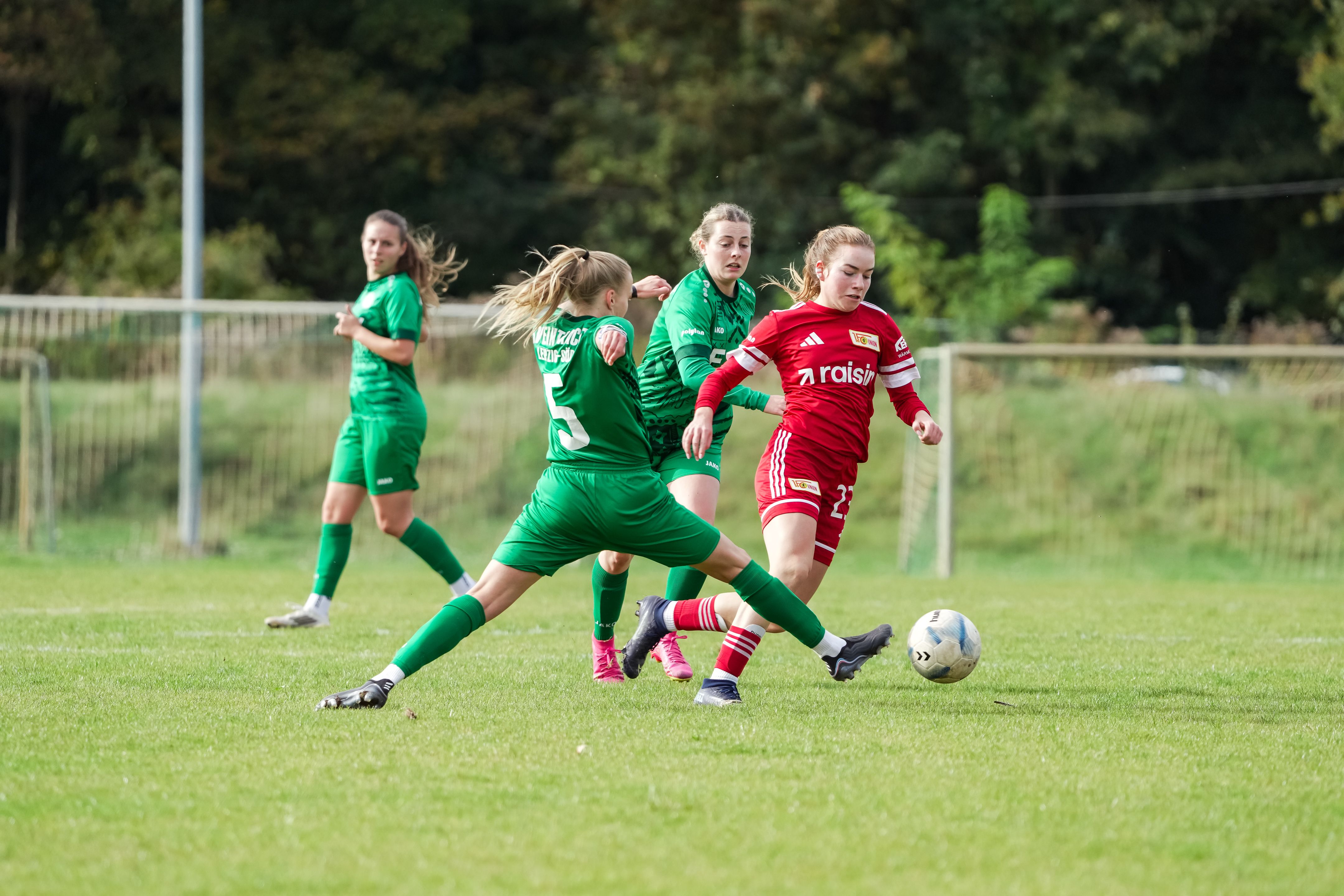 Zwei Fußballspielerinnen in grün und rot kämpfen um den Ball auf dem Spielfeld, während zwei weitere Spielerinnen im Hintergrund stehen.