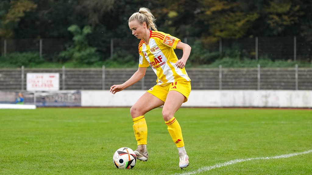 Fußballspielerin in gelb-weißem Trikot dribbelt einen Ball auf einem Rasenfeld.