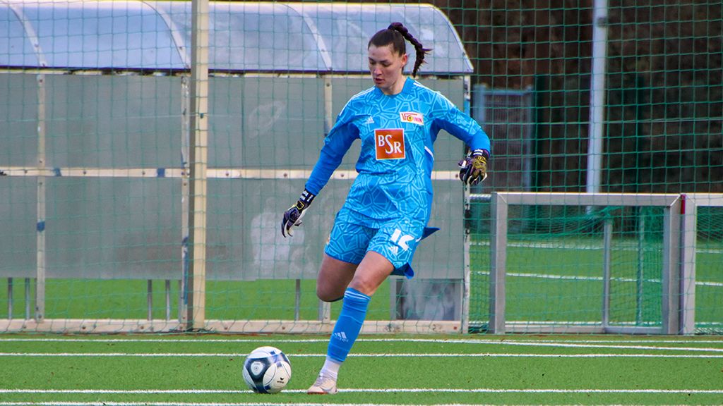A female goalkeeper in blue gear stands on the field, preparing to play the ball.