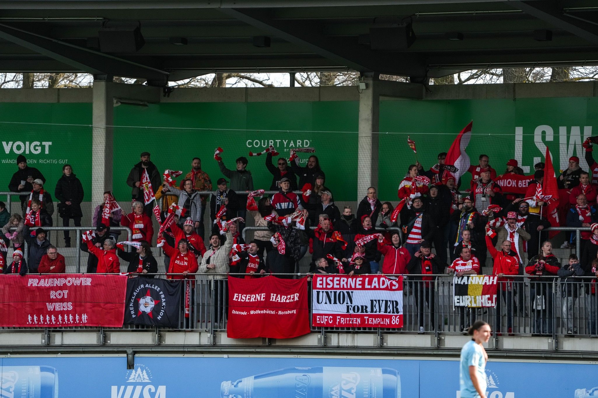 Fans stehen auf der Tribüne, halten Schals und Fahnen hoch, während sie ihr Team unterstützen. Banner im Vordergrund.