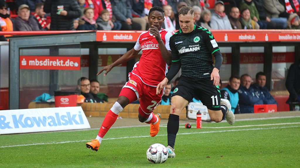 Two football players face each other while the ball is played between them in the stadium. Spectators in the background.