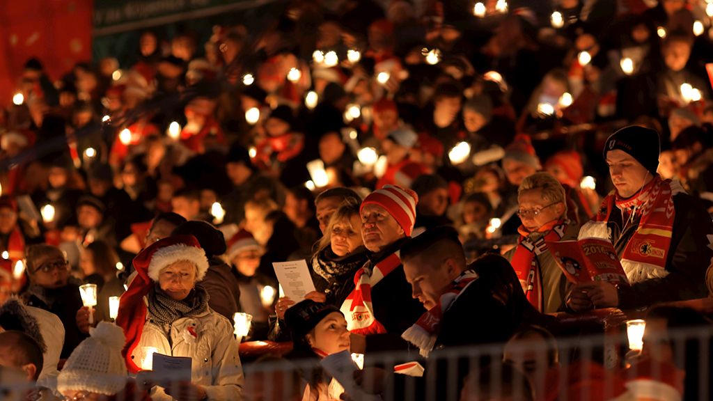 Eine Menschenmenge hält Kerzen in der Dunkelheit, viele tragen Weihnachtsmützen und lesen aus einem Liedblatt.