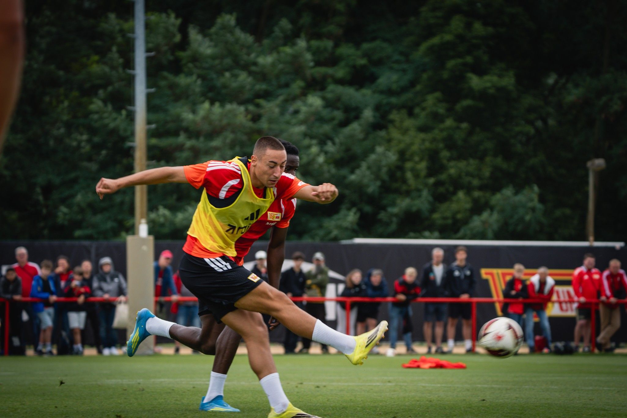 Ein Fußballspieler in gelber Trainingsweste schießt den Ball während eines Trainings auf dem Platz. Zuschauer sind im Hintergrund.