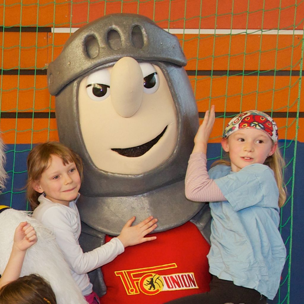 Ein maskottchenähnlicher Ritter mit zwei Kindern, die an ihm stehen und lachen, vor einer farbigen Wand aus Holzlatten.