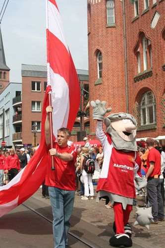 Ein junger Mann hält eine große rote und weiße Fahne, während ein Maskottchen neben ihm läuft. Im Hintergrund eine Menschenmenge.