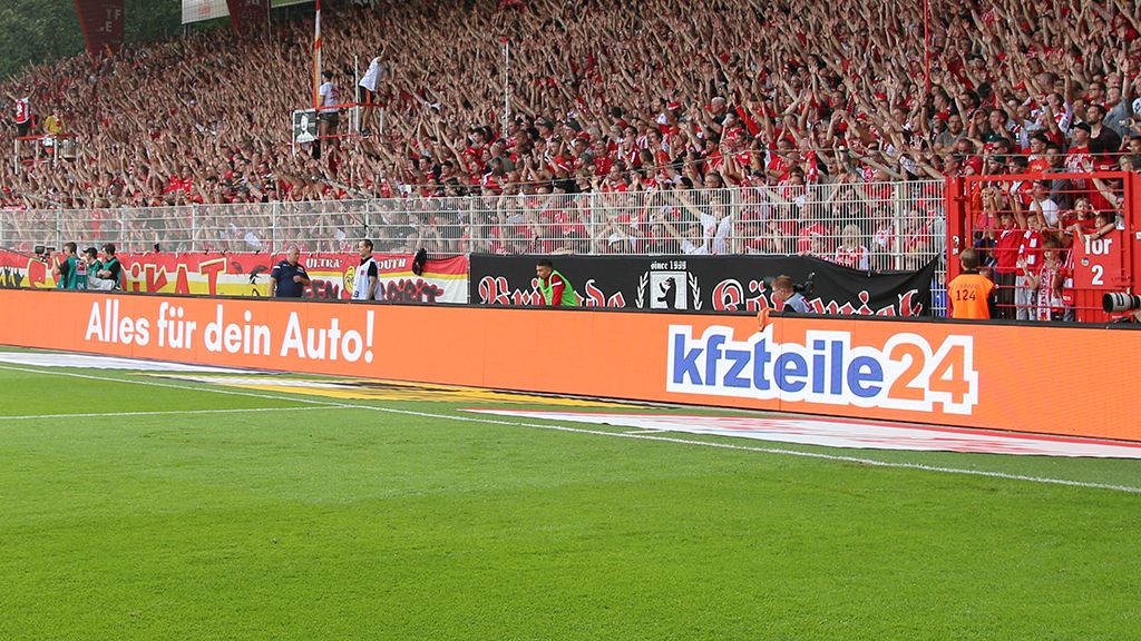 Ein Stadion mit jubelnden Fans, die rot gekleidete Plätze einnehmen, und Werbebanner am Spielfeldrand.